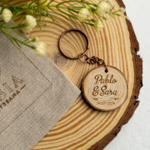 Llavero rústico de rodaja de madera para boda, personalizado con los nombres Pablo y Sara y la fecha, junto a una bolsa de lino y flores.