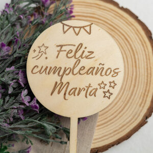 Topper de madera para tarta, personalizado con el grabado a láser "Feliz cumpleaños Marta", presentado junto a su bolsa de lino sobre una rodaja de madera.