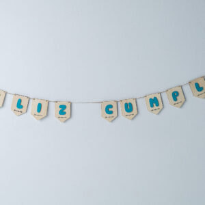 Guirnalda de madera para cumpleaños con banderines que forman la frase "FELIZ CUMPLE", con letras 3D de color turquesa, colgada en una pared blanca.