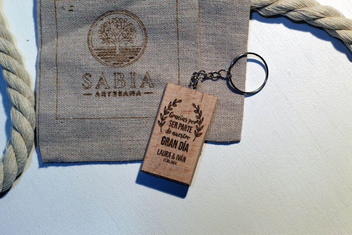 Llavero de madera personalizado con mensaje de agradecimiento para boda, presentado junto a su bolsa de tela de lino con el logo de Sabia Artesana.