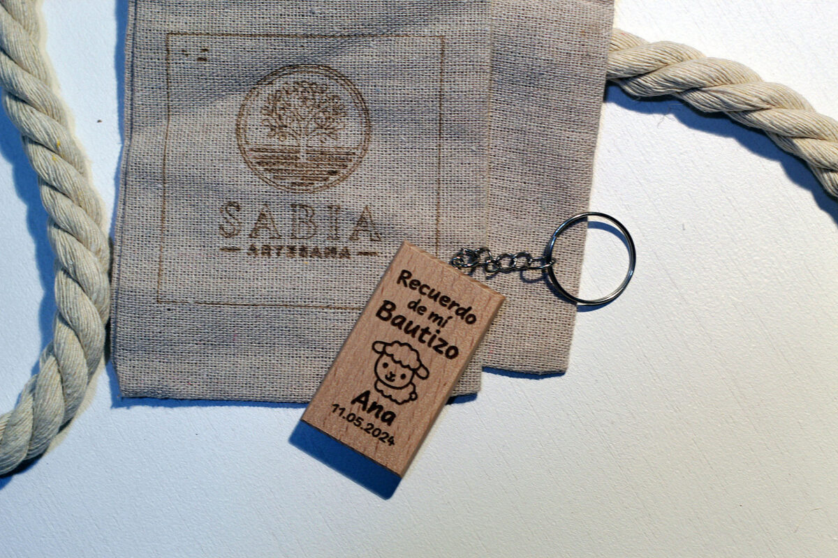 Llavero de madera para recuerdo de bautizo, grabado con una ovejita y el nombre Ana, mostrado junto a su bolsa de presentación de lino.