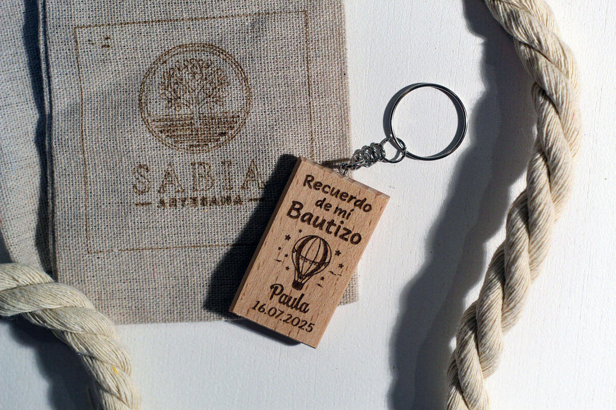 Recuerdo de bautizo: Llavero de madera rectangular personalizado con un globo y el nombre Paula, fotografiado junto a su bolsa de presentación.