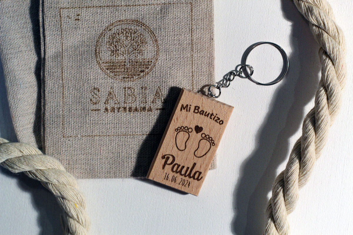 Llavero de madera rectangular para recuerdo de bautizo, grabado con unas huellas de pies de bebé y el nombre Paula, fotografiado junto a la bolsa de presentación de Sabia Artesana.