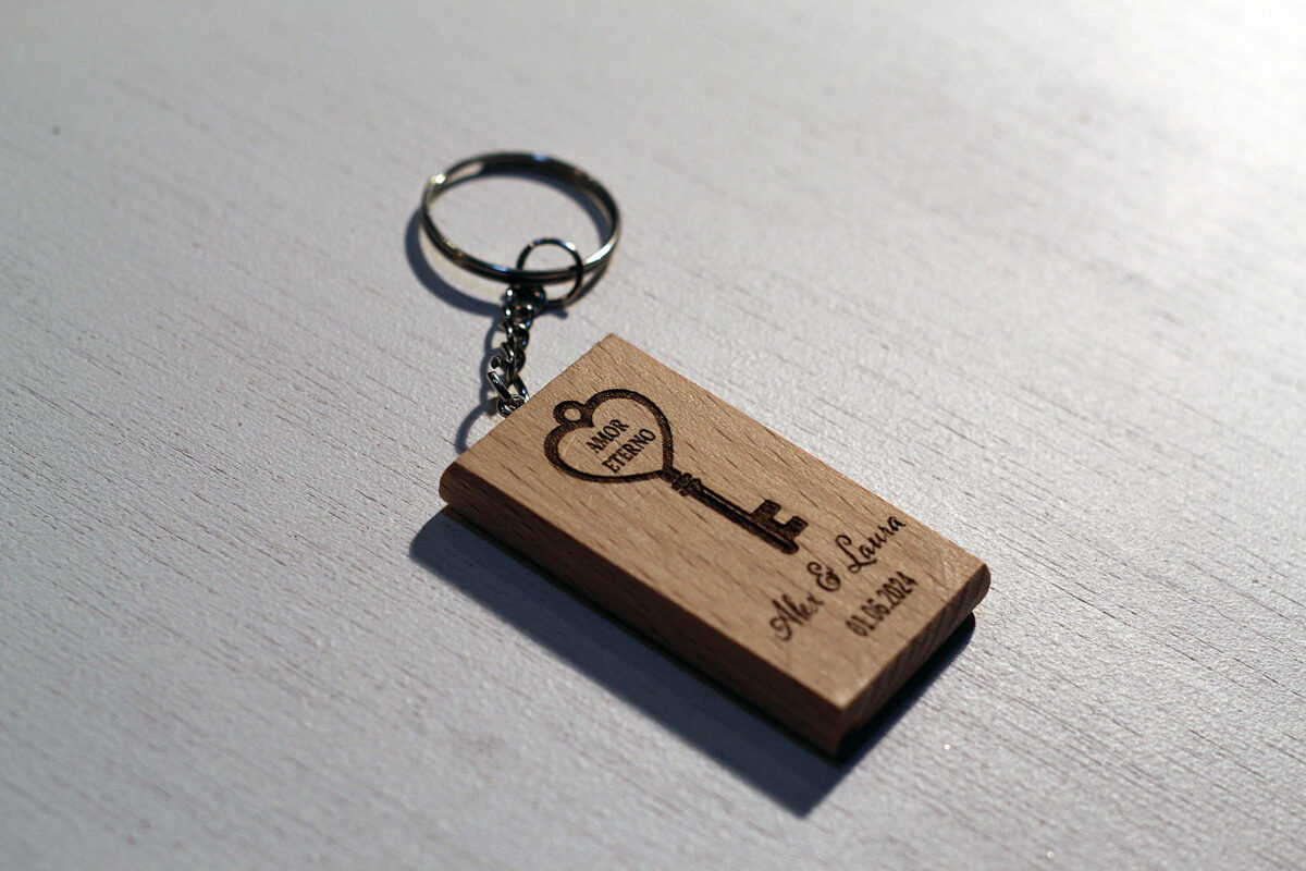 Detalle de boda original: Llavero de madera rectangular personalizado con los nombres Alex y Laura y un grabado de una llave con corazón y el texto Amor Eterno.